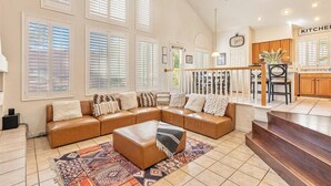 Un salon spacieux avec un grand canapé d’angle, un tapis à motifs et un coin cuisine doté de meubles en bois et d’une horloge au mur.