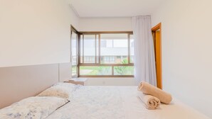 A hotel room with a bed, a nightstand, a window with curtains, and a view of greenery outside. - IT29 Excelente ap 3 Quartos 50m da Praia (Camaçari)