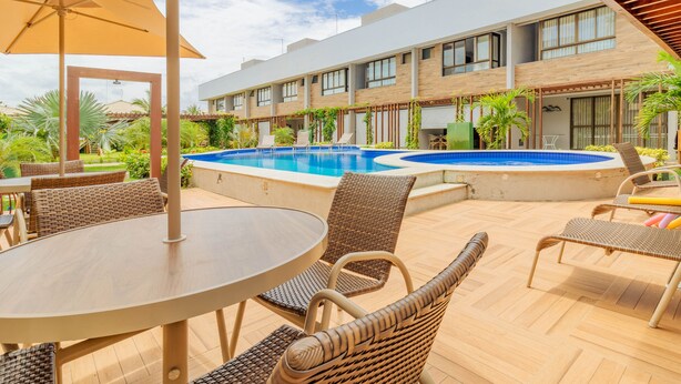 An outdoor pool area with lounge chairs and a table, surrounded by a modern building with large windows and a covered patio. - IT29 Excelente ap 3 Quartos 50m da Praia (Camaçari)