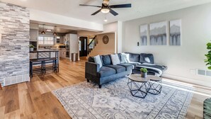 A spacious living area with a gray sectional sofa, a coffee table, and a patterned rug. A kitchen is visible in the background, and there is a staircase leading to another level. - Modern 5BR w/ Pool, Cinema & Game Room (Dallas)