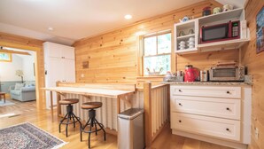 A kitchen with wooden walls, a wooden table, bar stools, a trash bin, a microwave, a toaster, and a window.