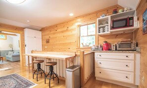 Eine Küche mit Holzwänden, einem Holztisch, Barhocker, einem Mülleimer, einer Mikrowelle, einem Toaster und einem Fenster.