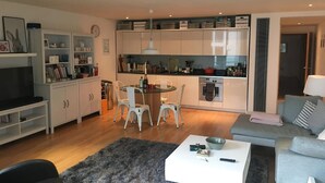 Un salon moderne avec un canapé gris, une table basse et un téléviseur. On y trouve également un coin repas avec une table et des chaises, ainsi qu'une cuisine dotée de meubles blancs et d'appareils électroménagers.