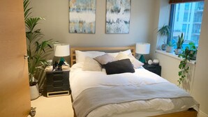 Une chambre à coucher avec un lit en bois, deux tables de chevet avec des lampes, une fenêtre ornée de plantes et des œuvres d'art abstraites accrochées au mur.