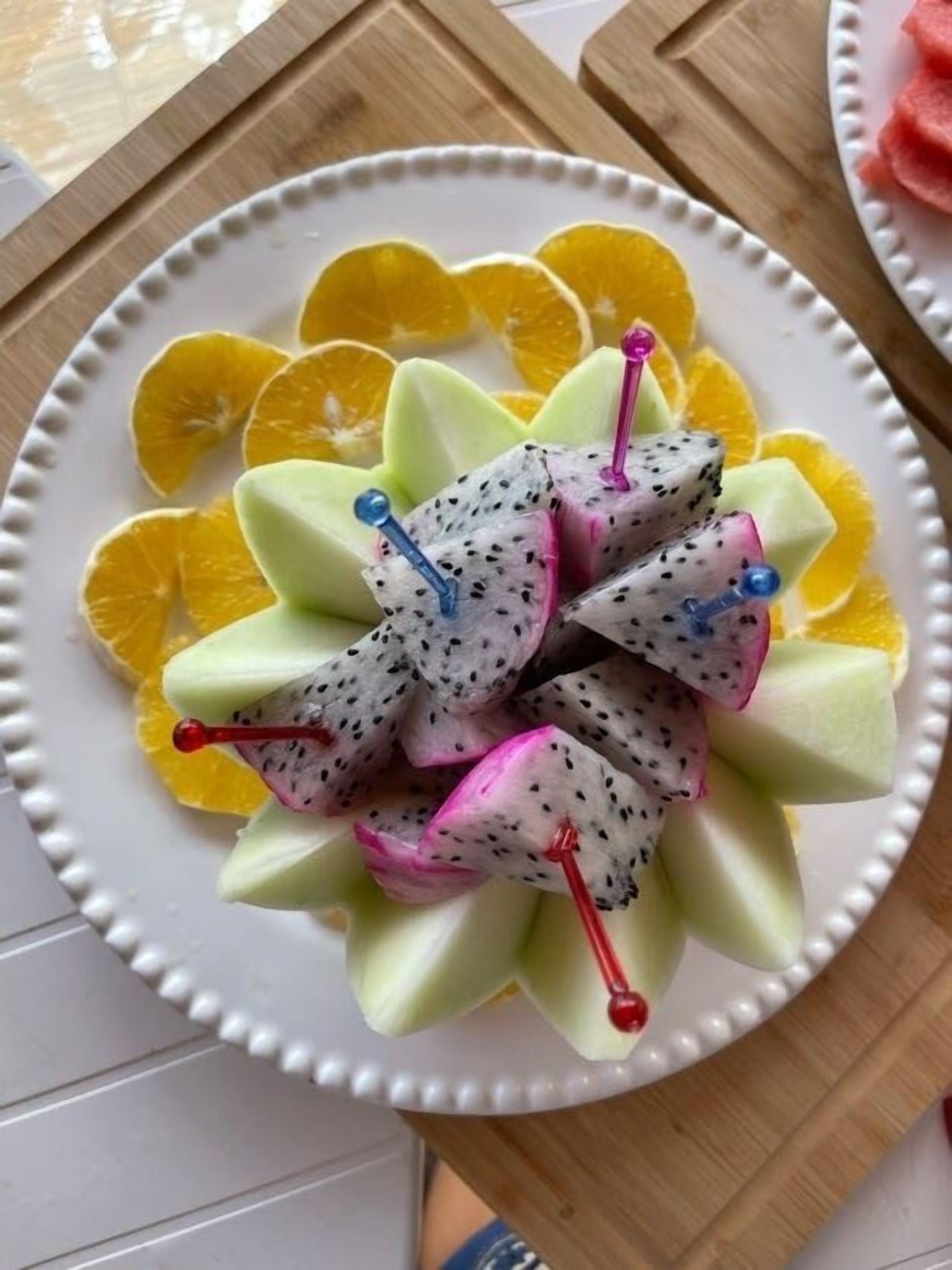 A plate of dragon fruit slices arranged on a white plate with a decorative edge, surrounded by orange slices.