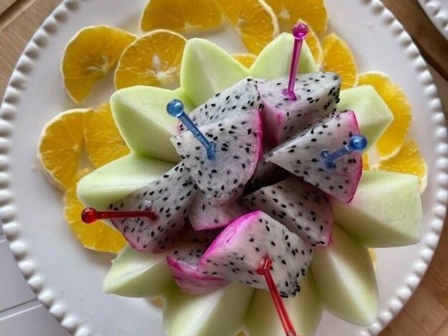 A plate of dragon fruit slices arranged on a white plate with a decorative edge, surrounded by orange slices.