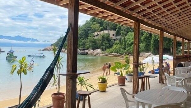 A covered outdoor seating area with tables and chairs, overlooking a beach and water.