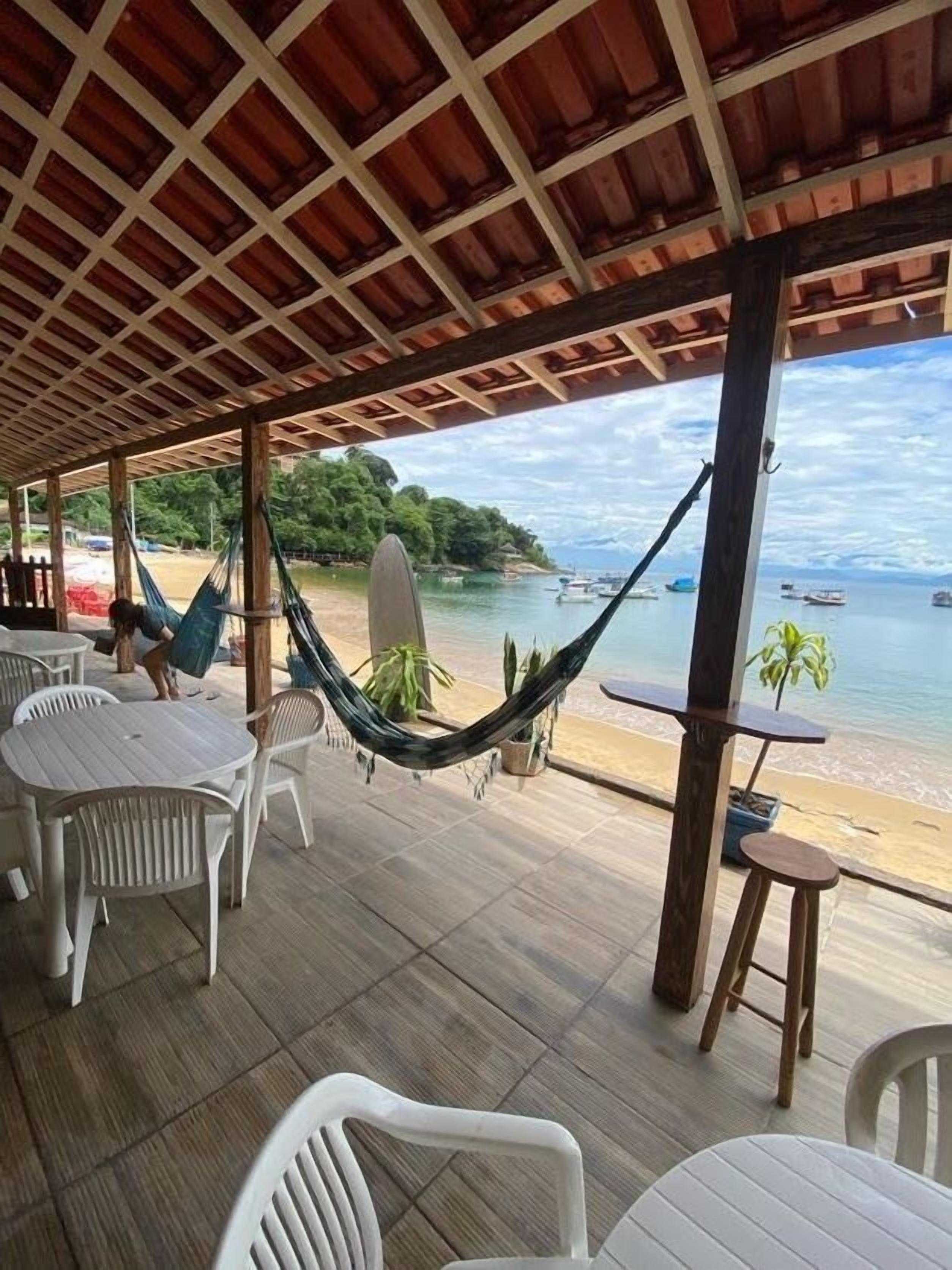 A covered outdoor seating area with tables and chairs, a hammock, and a view of the beach and boats.