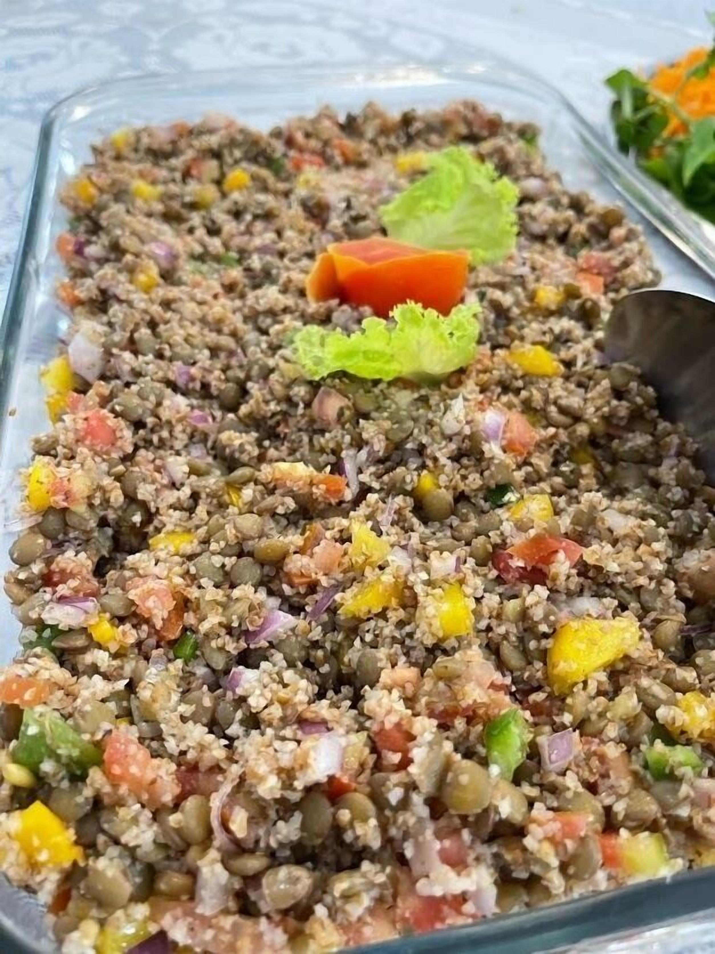 A glass container filled with a grain-based salad, possibly containing quinoa, mixed with various vegetables and garnished with a leaf of lettuce.