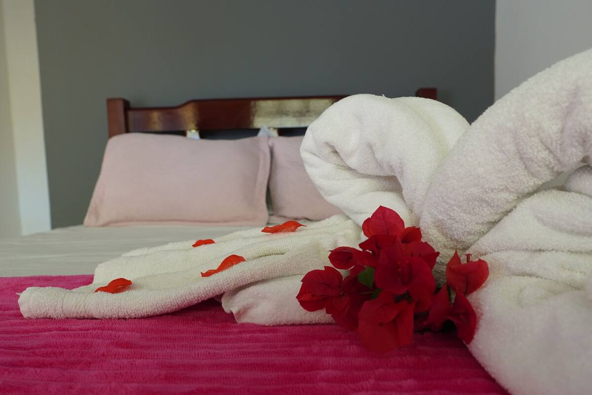A neatly made bed with white and red towels, a pink blanket, and red flowers on a pink bedspread.
