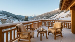 A wooden balcony with two chairs and a small table, overlooking a snow-covered village and mountains.