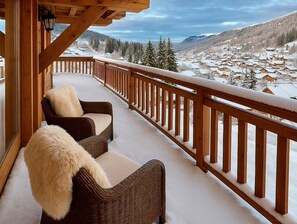 A wooden balcony with two wicker chairs and a fur throw, overlooking a snowy village and mountains.