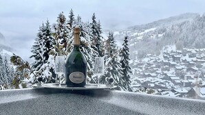 A bottle of Ruinart champagne on a balcony with snow-covered trees and a snowy village in the background.