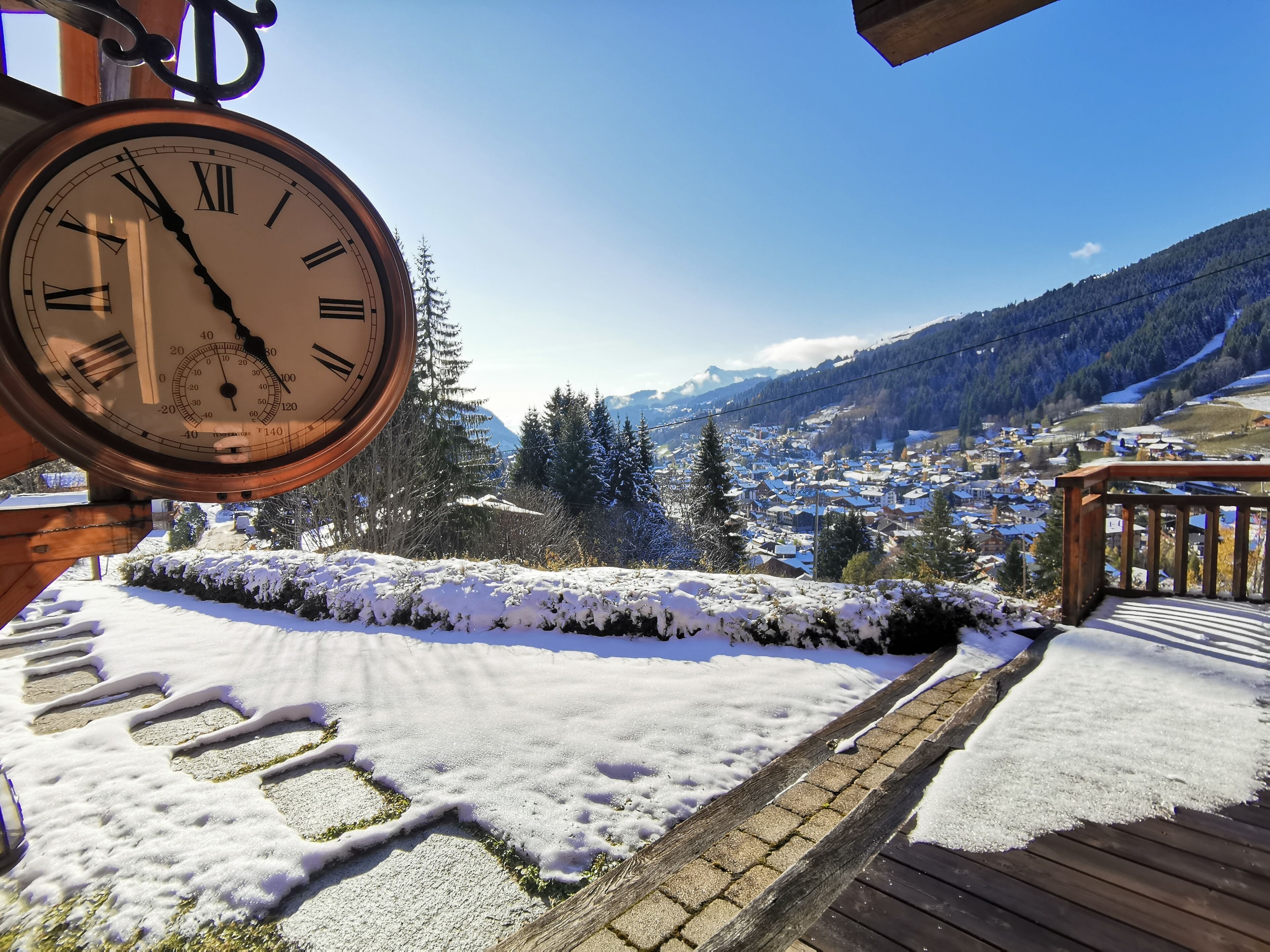 A clock with Roman numerals, a wooden deck, and a snowy landscape with a town in the background.