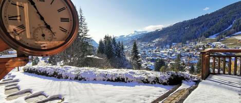 A clock with Roman numerals, a wooden deck, and a snowy landscape with a town in the background.