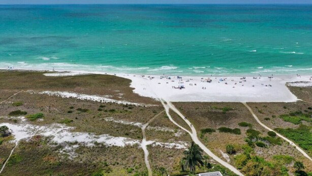 Luchtbeeld van een strand met een helder pad dat naar de kust leidt, omgeven door begroeiing en enkele verspreide gebouwen.