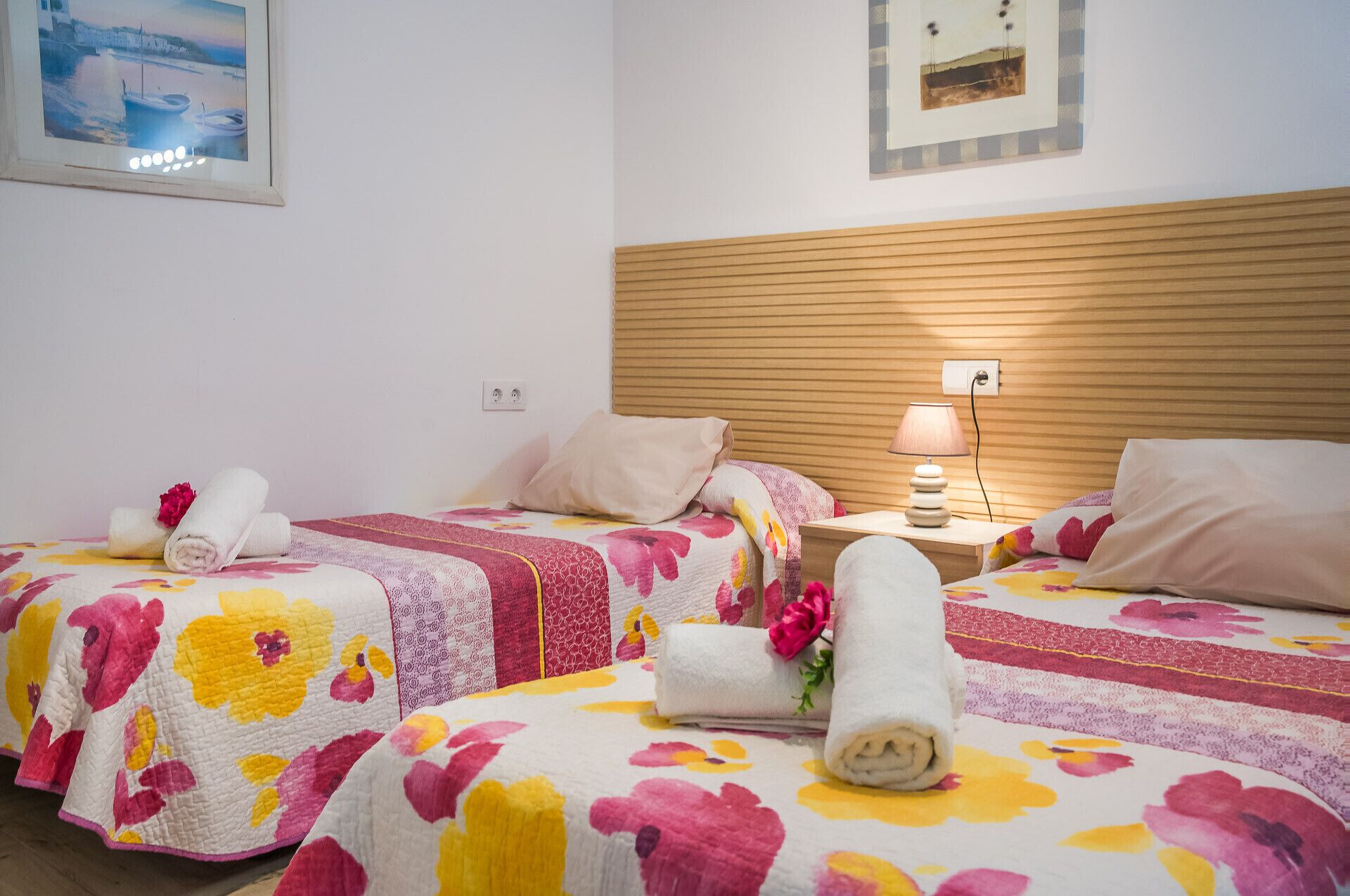 Habitació d'hotel amb dos llits, roba de llit floral, un panell de fusta al capçal, una nit de nit amb una làmpada, i un quadre amb marc a la paret.