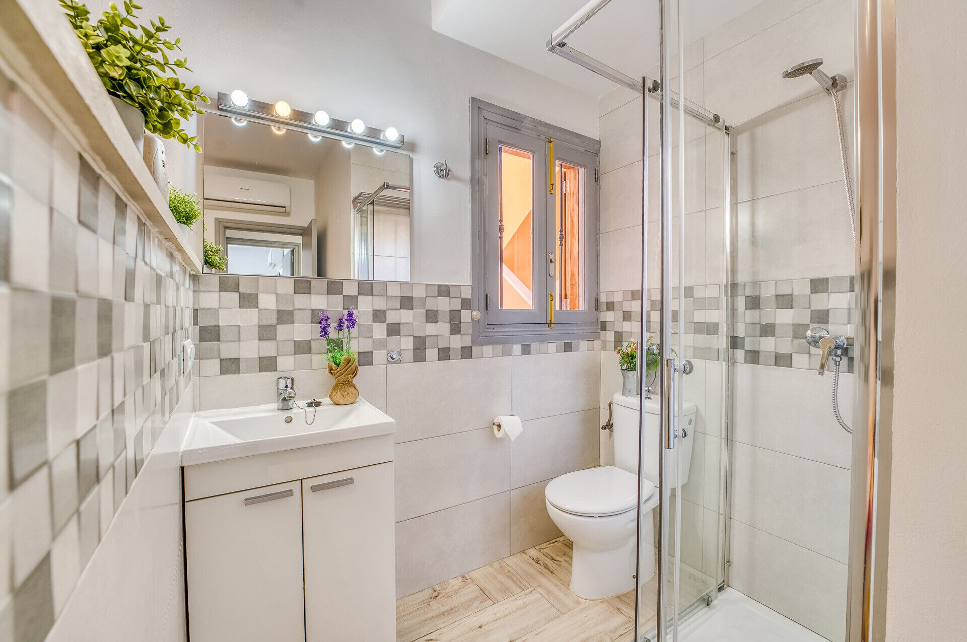 A modern bathroom with a glass shower enclosure, a vanity with a mirror and lights, a toilet, and a window with a view.