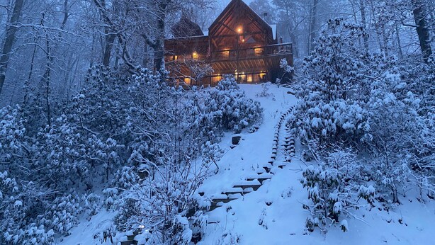 Una cabaña de troncos con una chimenea central, rodeada de árboles cubiertos de nieve y un sendero nevado que lleva a la entrada.