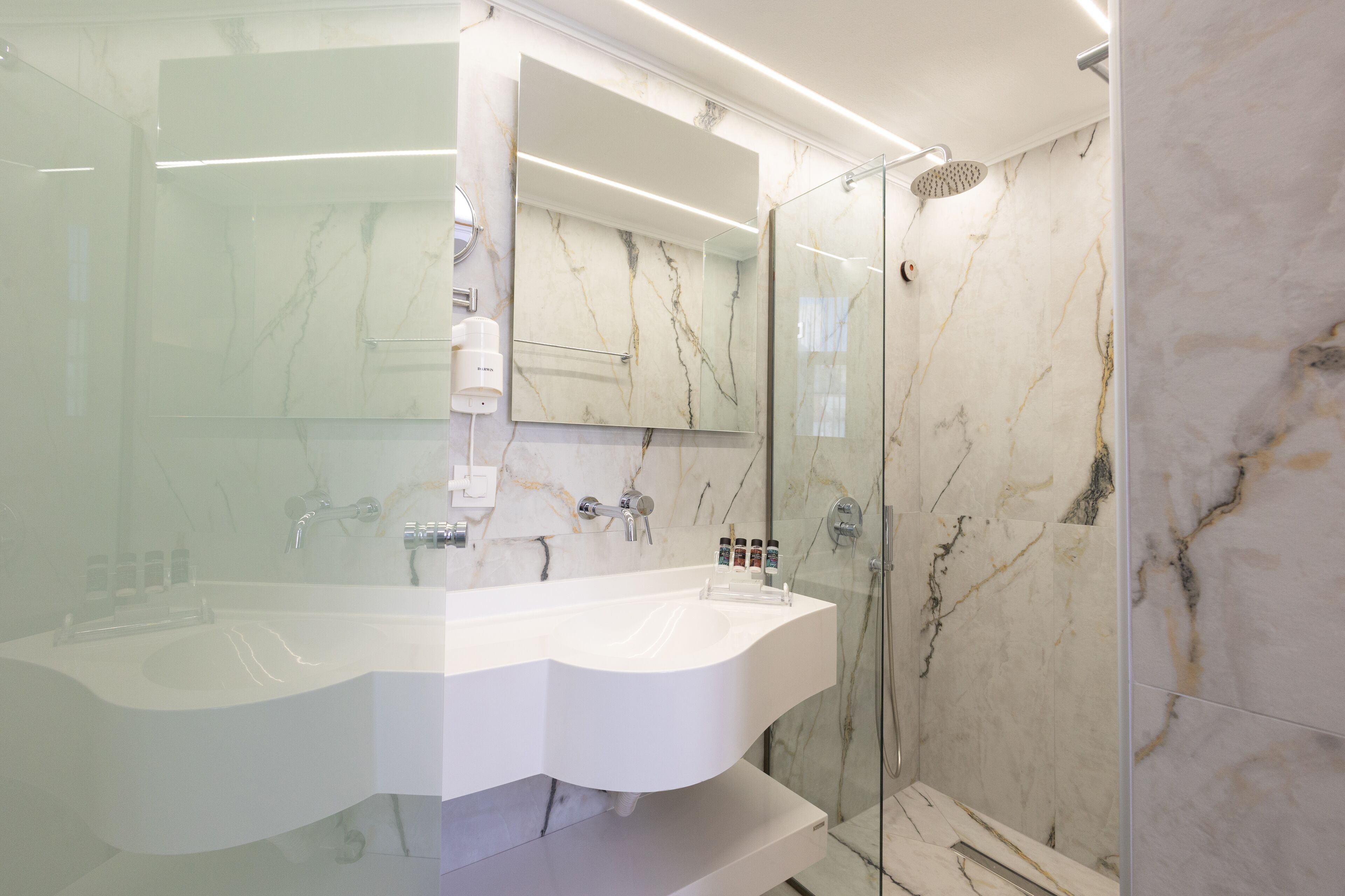 A modern bathroom with a large white sink, a glass-enclosed shower, and marble walls.