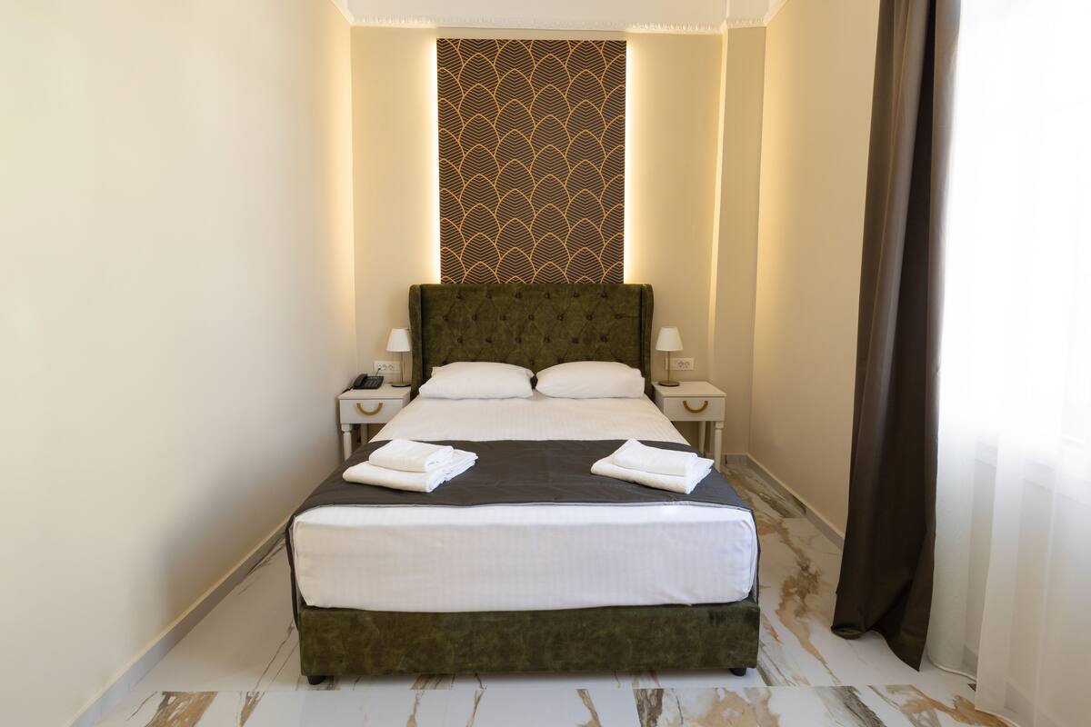 Una cama bien hecha con sábanas blancas, dos mesitas de noche con lámparas y un papel tapiz estampado detrás de la cama.