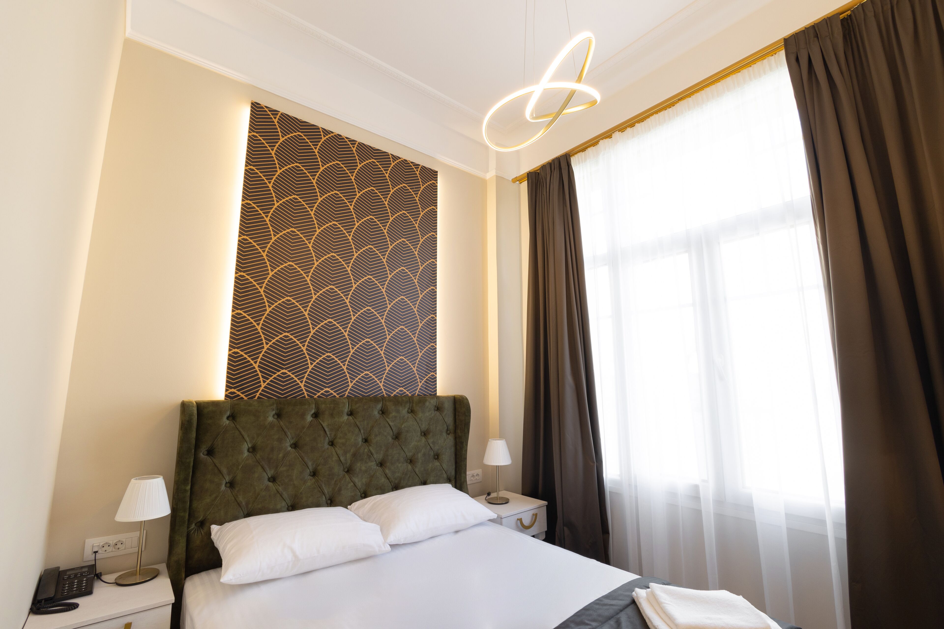 Une chambre d’hôtel avec un grand lit, une table de chevet avec une lampe, un mur orné d’un motif géométrique et une fenêtre avec des rideaux.