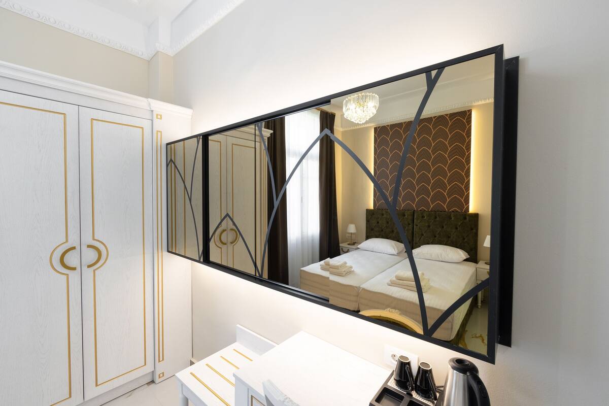 Habitación de hotel con un espejo grande, una cama con ropa de cama impecable, una mesita de noche con lámpara y un armario con detalles dorados.
