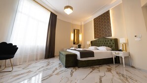 Habitación de hotel con una cama grande, un banco tapizado de verde, una mesita de noche blanca, una silla negra y un ventanal con cortinas traslúcidas.