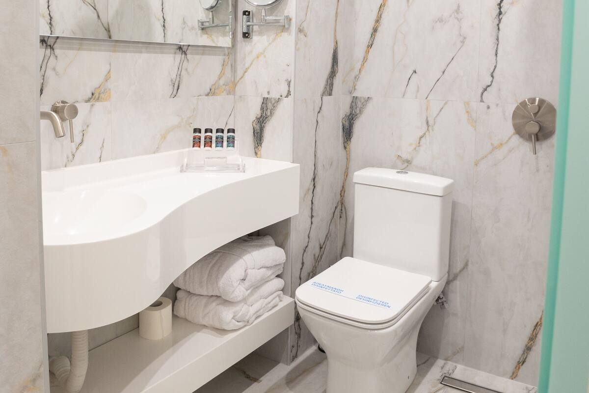 Un baño con lavamanos blanco, espejo, inodoro, paredes de mármol y ducha.