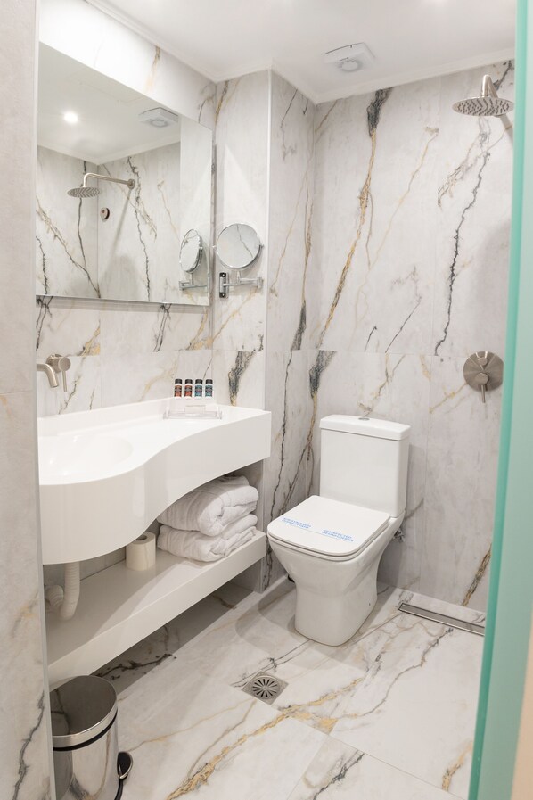 Un baño con lavamanos blanco, espejo, inodoro, paredes de mármol y ducha.