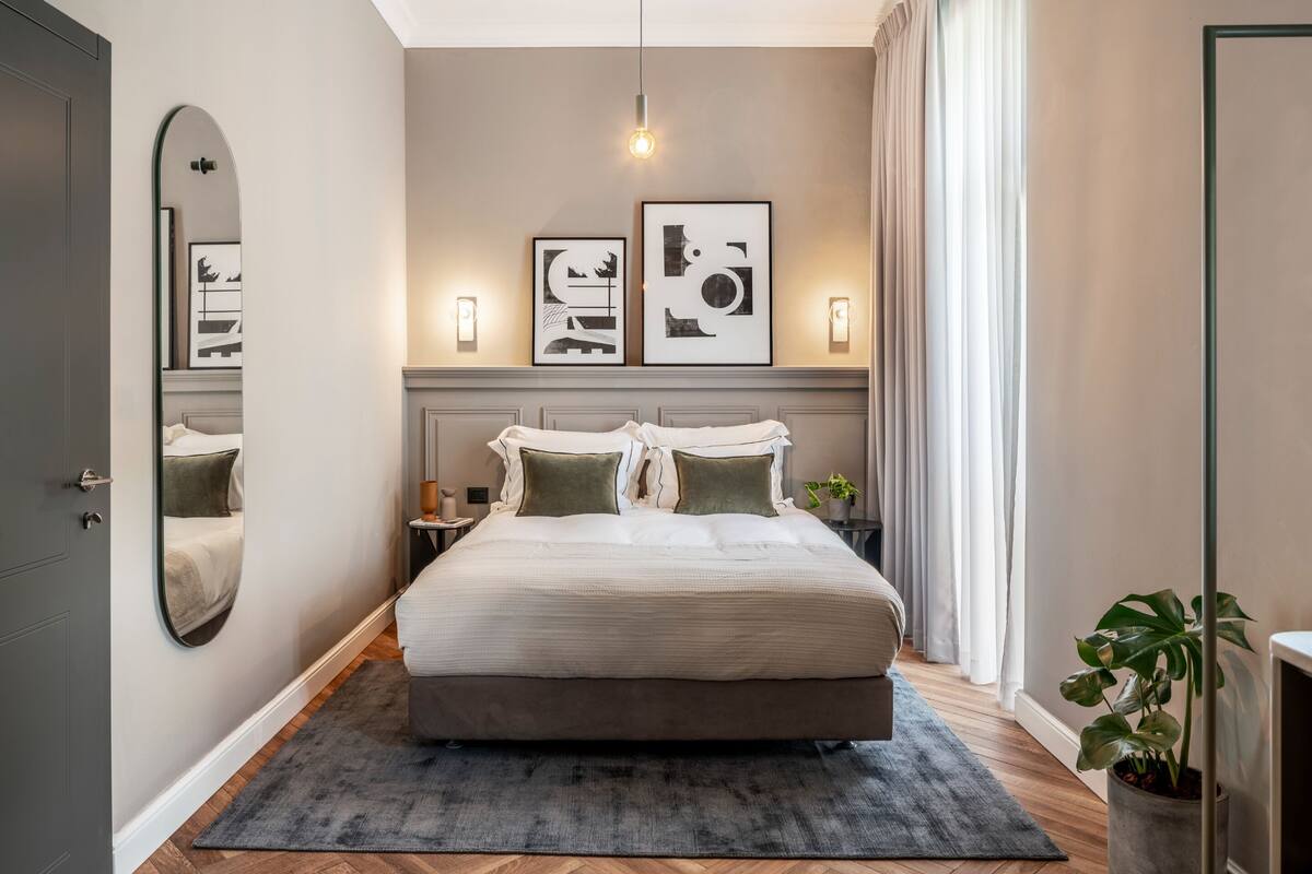 A modern bedroom with a large bed, a mirror, framed artwork, and a potted plant.