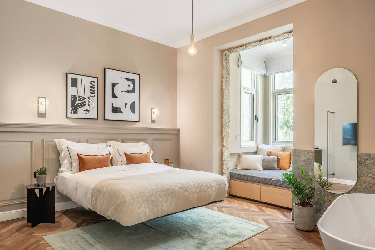 A bedroom with a large bed, a window seat, a bathroom with a bathtub, and two wall-mounted light fixtures.