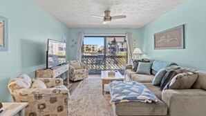 Ein Wohnzimmer mit Sofa, Sessel und Couchtisch, einem Deckenventilator und Blick durch eine Glastür nach draußen.