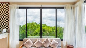 Quarto com uma janela grande, um sofá com almofadas e um banquinho de madeira.