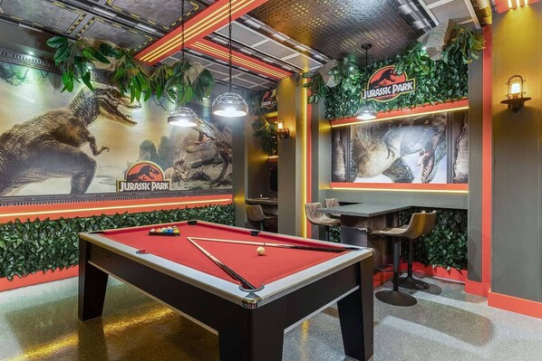 A pool table with a red felt surface, surrounded by walls decorated with dinosaur murals and greenery.