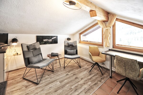 A cozy loft bedroom with a large window, wooden beams, and modern furniture including a desk and two chairs.