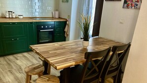 A dining area with a wooden table and four chairs, a potted plant, and a green kitchen in the background.