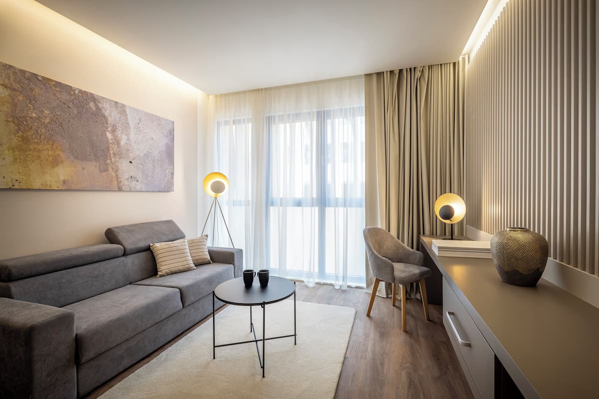 Een moderne woonkamer met een grijze bank, een kleine ronde tafel en een stoel. Aan de muur hangt een groot abstract schilderij, er staat een vloerlamp en een vaas op een bijzettafeltje.