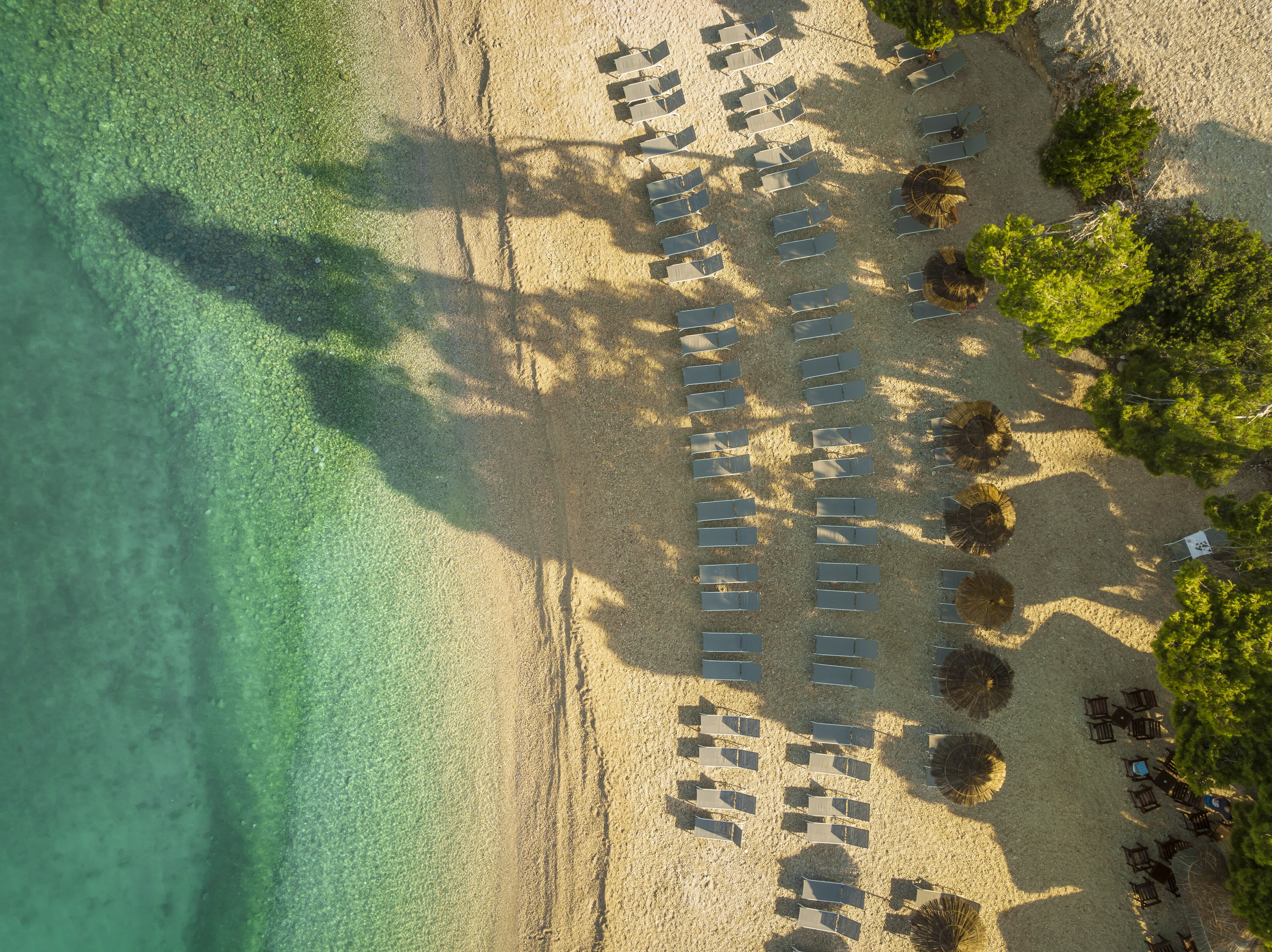 An aerial view of a beach with rows of sun loungers and umbrellas, clear turquoise water, and lush greenery.