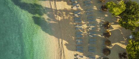 An aerial view of a beach with rows of sun loungers and umbrellas, clear turquoise water, and lush greenery.