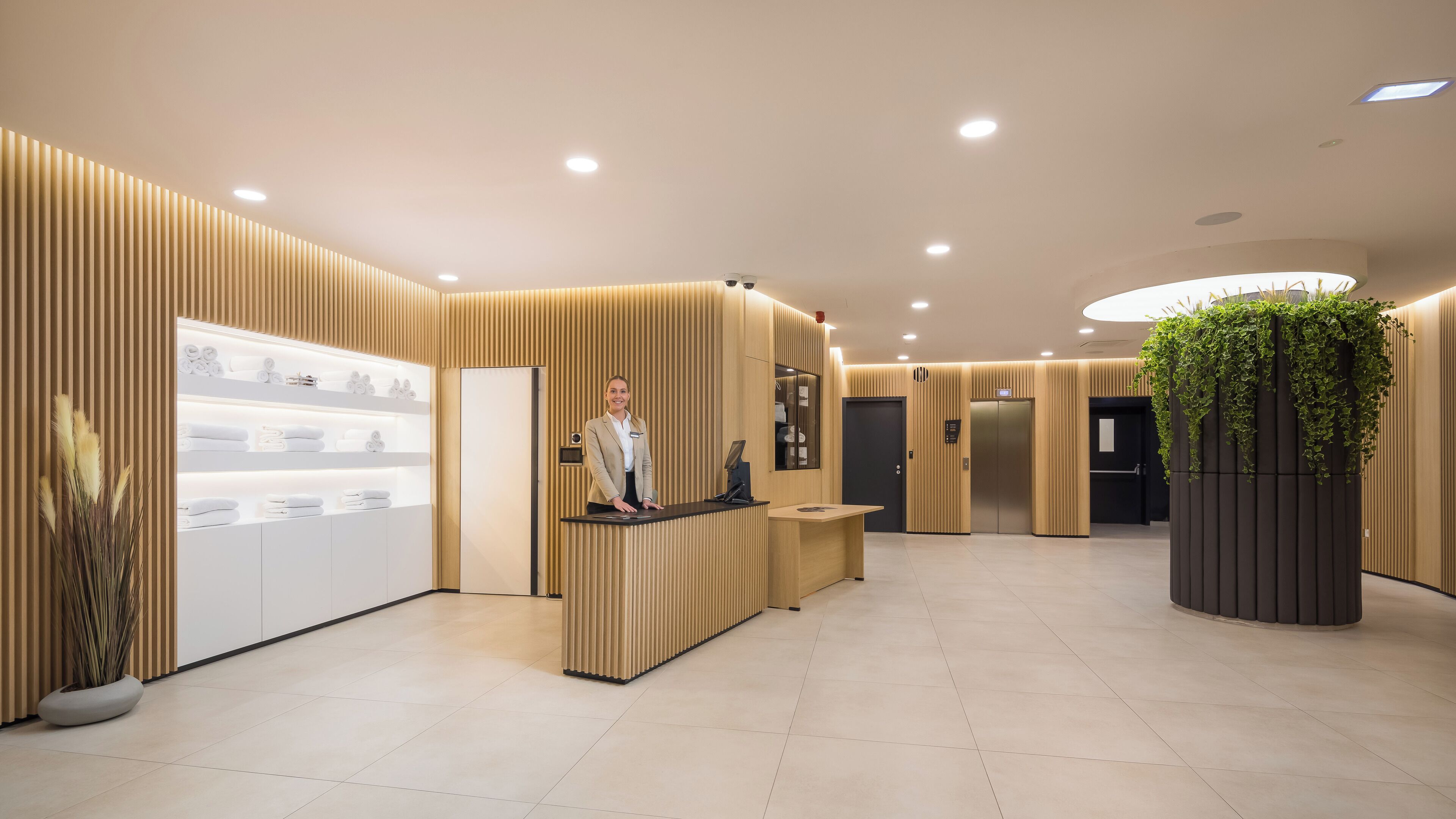 Una zona de recepción moderna con recepcionista, mostrador de recepción, planta y estante con toallas.