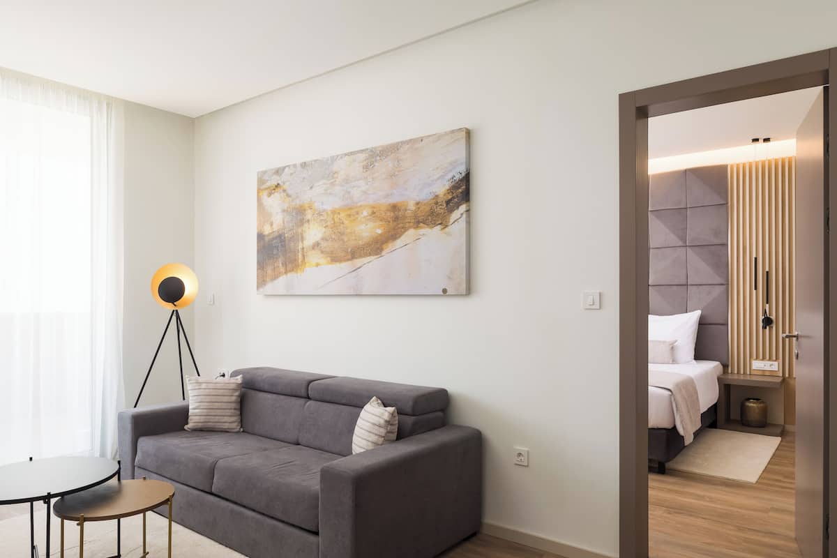 Een moderne hotelkamer met een grijze bank, een ronde salontafel, een vloerlamp en een groot abstract schilderij aan de muur.