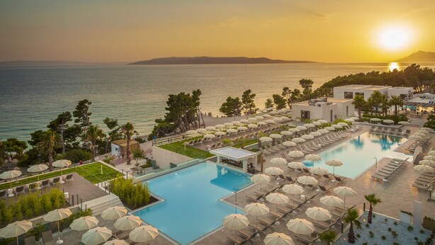 Een resort met een groot zwembad, ligbedden, parasols en uitzicht op zee bij zonsondergang.