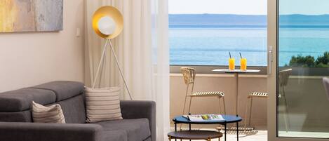 Un hotel moderno con un sofá gris, una mesita redonda con una revista, y vistas al mar desde un balcón.