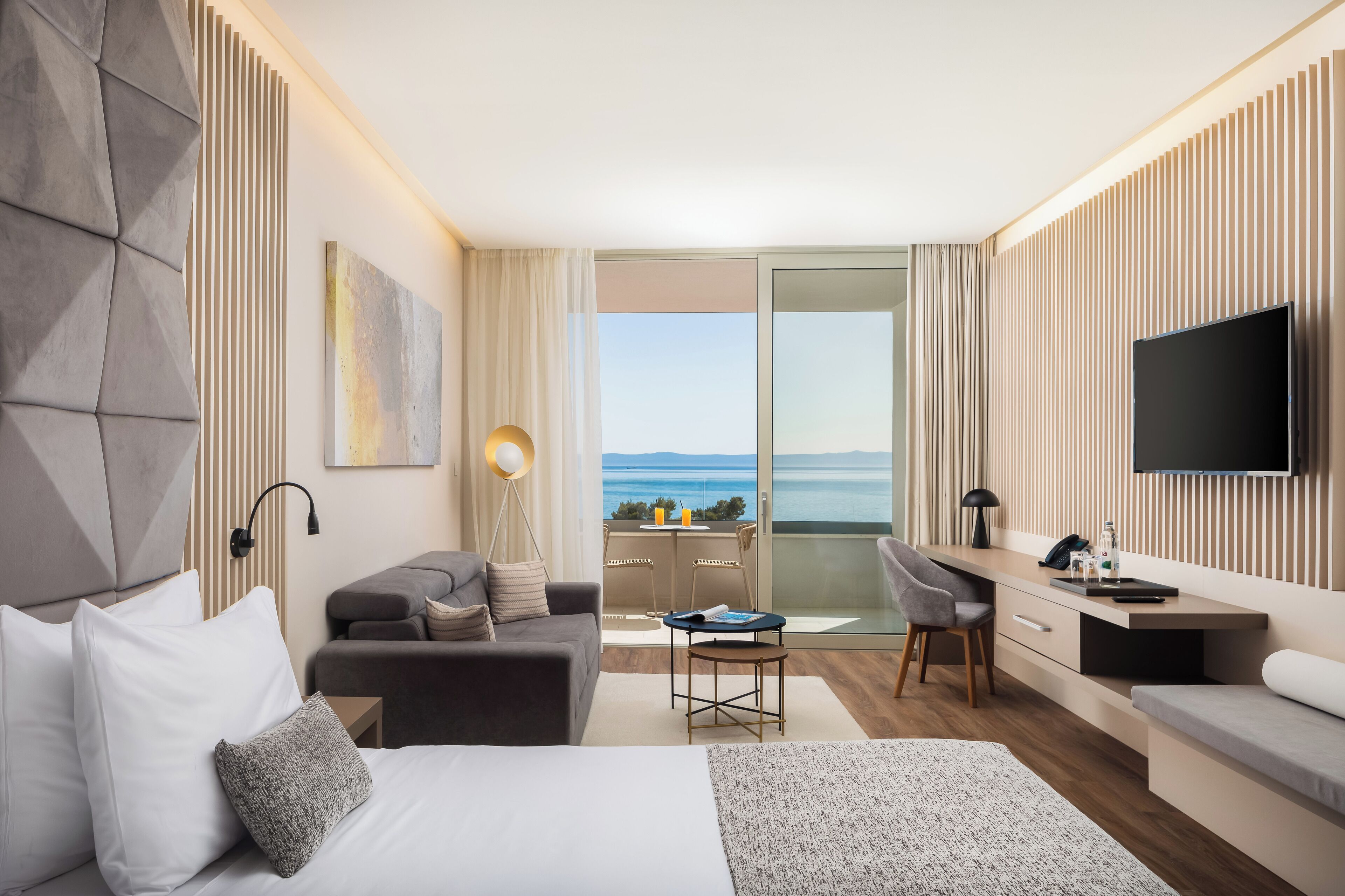Habitación de hotel moderna con cama, sofá, escritorio y televisor. Posee un balcón con vista al mar.