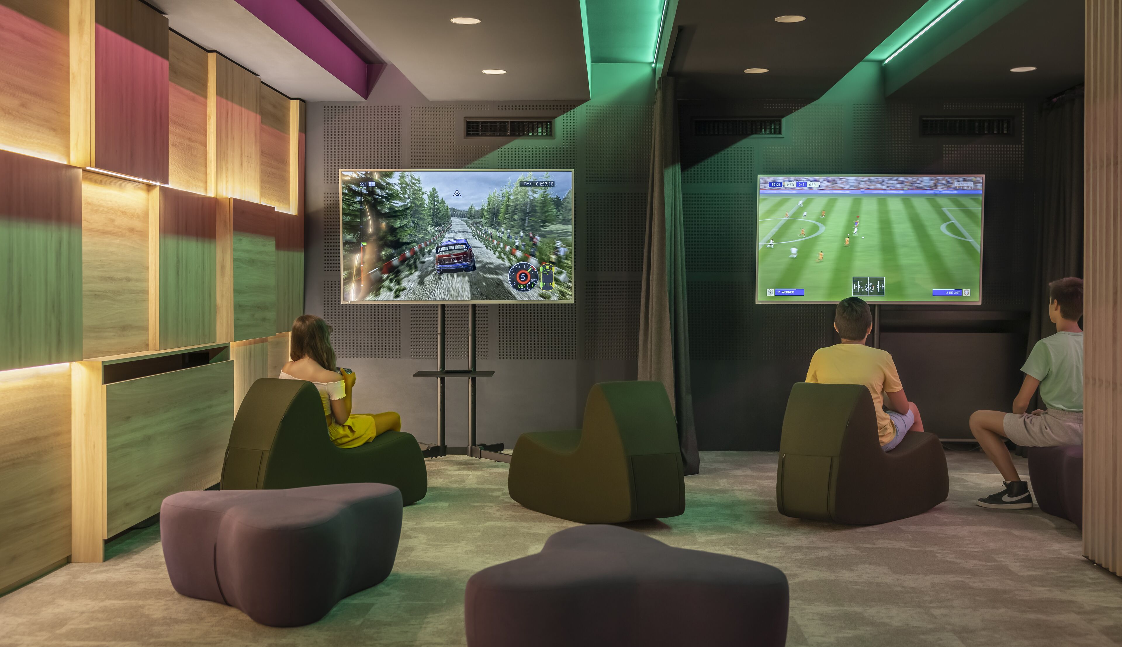 A modern gaming room with two large screens displaying racing and soccer games. There are comfortable seating areas and a wooden panel wall.
