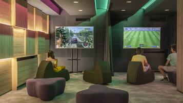 A modern gaming room with two large screens displaying racing and soccer games. There are comfortable seating areas and a wooden panel wall.