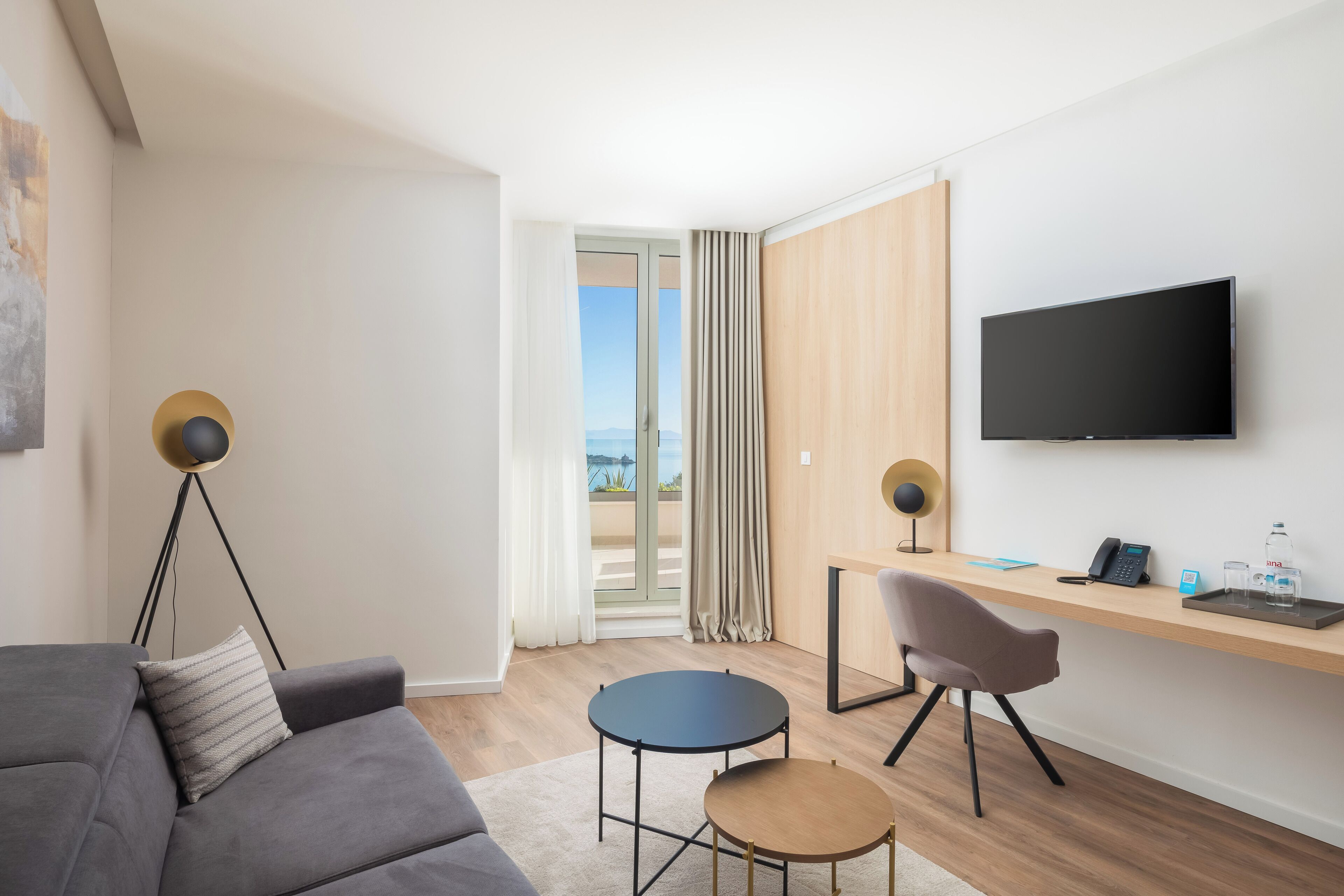 Una habitación de hotel moderna con un sofá gris, una pequeña mesa redonda, un escritorio con una silla, un televisor de pantalla plana y vistas al exterior a través de una puerta corredera.