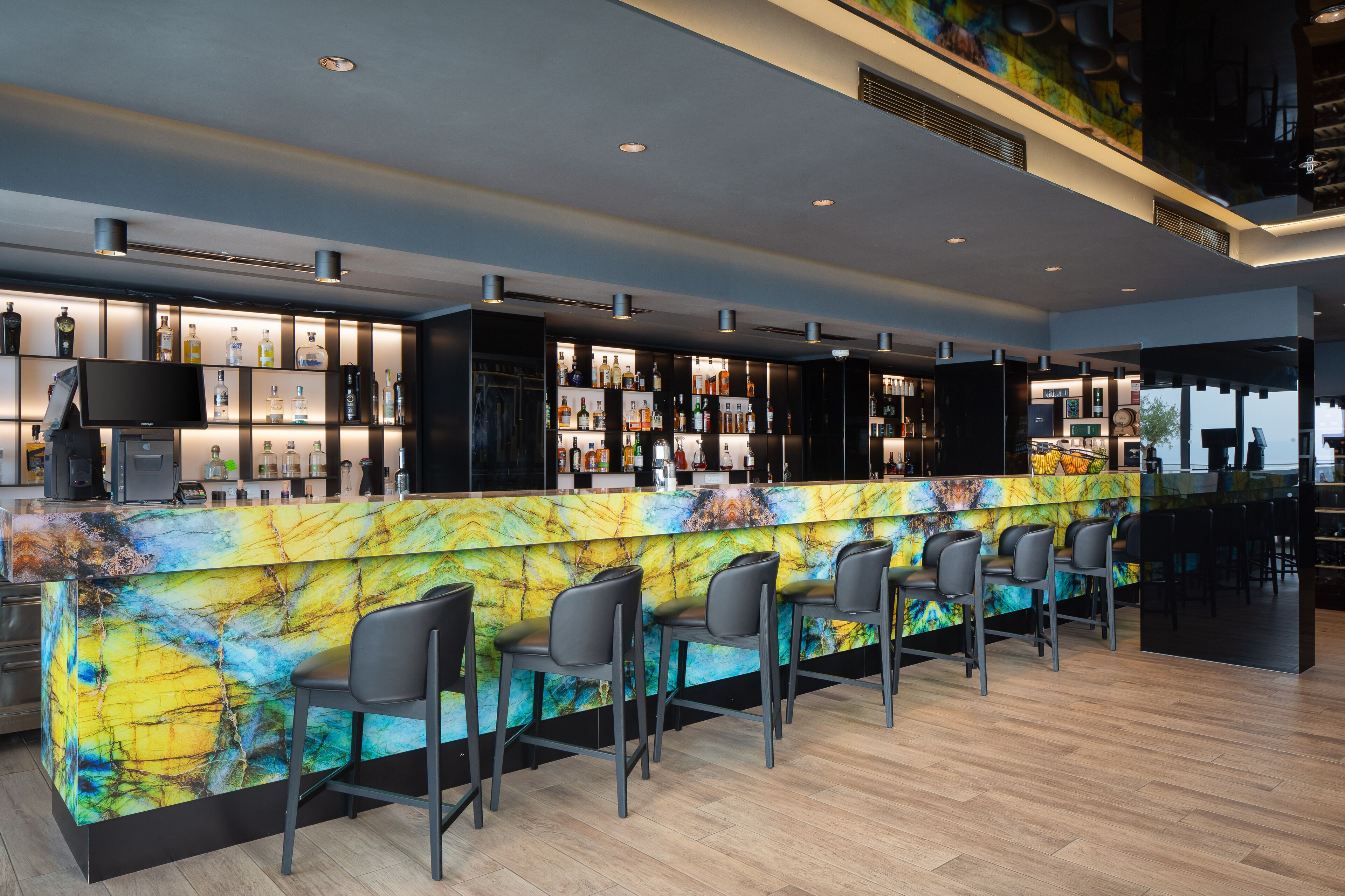 Zona de bar moderna con una encimera de mármol de colores, varios taburetes y una variedad de botellas y vasos en estanterías.