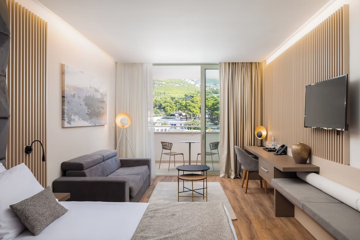 Een moderne hotelkamer met een bed, een bank, een bureau en een televisie. Er is een balkon met uitzicht op de stad en groen.